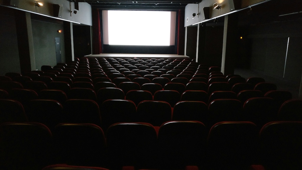 Salle de cinéma vide avec écran blanc lumineux, sièges rouges.